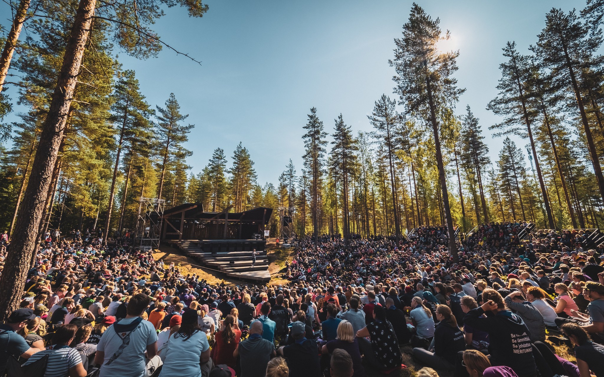 Aurinkoinen metsämaisema, jossa on paljon ihmisiä kuuntelemassa puhujaa Johtajatulien lavalla.